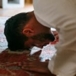 A person in traditional attire bows in prayer on a patterned rug within a room.